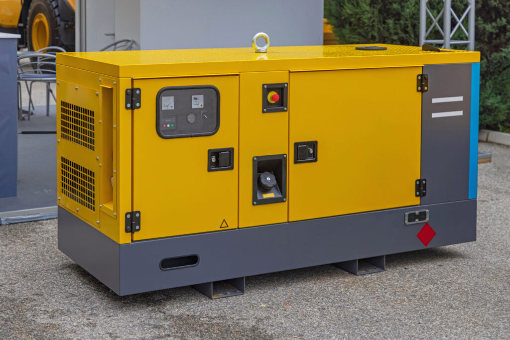 A portable generator with a bright yellow and gray design is situated outdoors on a paved surface. The generator features control panels, ventilation grilles, and safety labels. It is equipped with various access points for operation and maintenance. The background includes other industrial equipment, indicating the generator's use in a commercial or construction setting.