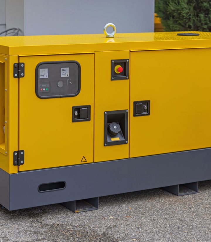 A portable generator with a bright yellow and gray design is situated outdoors on a paved surface. The generator features control panels, ventilation grilles, and safety labels. It is equipped with various access points for operation and maintenance. The background includes other industrial equipment, indicating the generator's use in a commercial or construction setting.