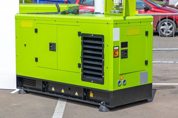 A bright green portable generator is situated on a paved surface, surrounded by parked cars. The generator features multiple access panels, ventilation grilles, and control buttons, including a prominent red emergency stop button. Tools and equipment are placed on top of the generator, indicating active use or maintenance. The vibrant color and compact design make it suitable for various applications, from industrial to event support.
