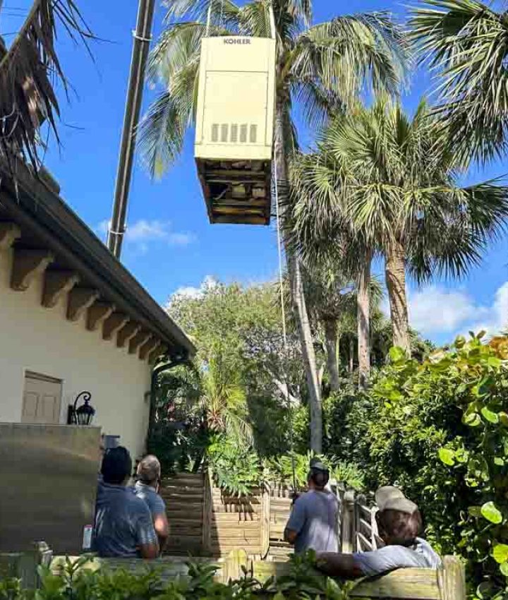 A crane is seen lifting a Kohler generator high above a residential area for installation. Workers on the ground are guiding the process, ensuring safe and precise placement. The setting is surrounded by lush tropical vegetation and palm trees, with a house partially visible in the background. The sky is clear and blue, highlighting a sunny day ideal for installation work.