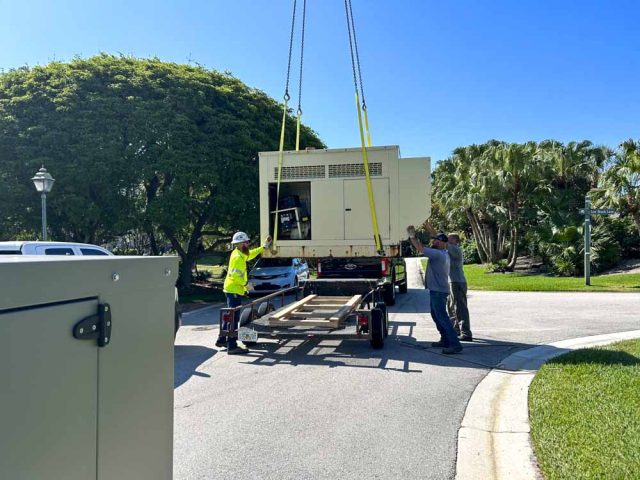 A crane is used to lift a large generator onto a trailer during an installation process. Workers are guiding the generator, ensuring it is properly positioned. The scene takes place in a residential area with lush green trees and a clear blue sky. Safety gear and equipment indicate a professional and secure operation.