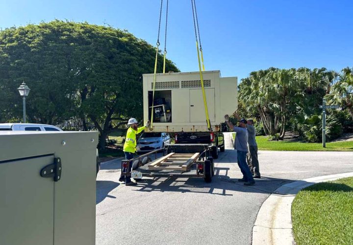 A crane is used to lift a large generator onto a trailer during an installation process. Workers are guiding the generator, ensuring it is properly positioned. The scene takes place in a residential area with lush green trees and a clear blue sky. Safety gear and equipment indicate a professional and secure operation.