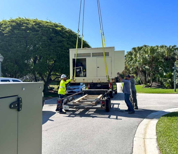 A crane is used to lift a large generator onto a trailer during an installation process. Workers are guiding the generator, ensuring it is properly positioned. The scene takes place in a residential area with lush green trees and a clear blue sky. Safety gear and equipment indicate a professional and secure operation.