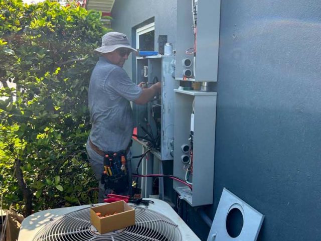 An electrical technician is installing and wiring electrical panels on the exterior wall of a house. The technician, wearing a hat and utility belt, is focused on the task. An air conditioning unit is positioned nearby, with various tools and equipment visible around the work area. The scene is surrounded by greenery, highlighting the residential nature of the installation.