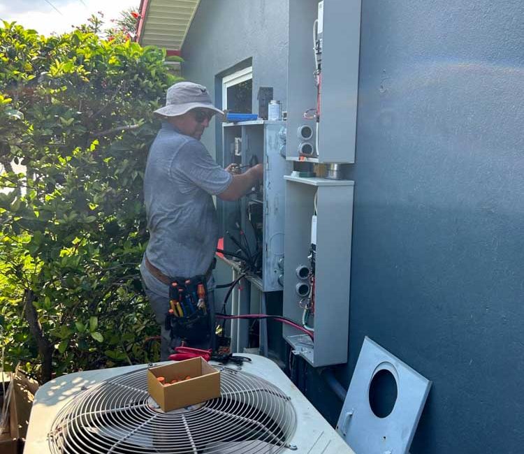 An electrical technician is installing and wiring electrical panels on the exterior wall of a house. The technician, wearing a hat and utility belt, is focused on the task. An air conditioning unit is positioned nearby, with various tools and equipment visible around the work area. The scene is surrounded by greenery, highlighting the residential nature of the installation.