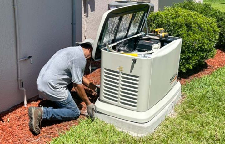 A residential electrician in Melbourne, FL, kneels beside a Generac backup generator, working on its installation. The generator's cover is open, exposing the internal components. The technician is positioned next to the house, with a gas meter visible nearby. The surrounding area is landscaped with mulch and bushes, indicating a residential setting. Tools and equipment are placed nearby, highlighting the active installation process.
