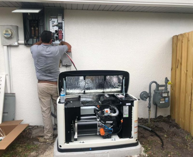 An electrical contractor in Melbourne, FL, is working on the installation of a generator outside a house. The generator cover is open, revealing its internal components. The contractor is connecting wires to an electrical panel mounted on the exterior wall. The scene includes utility meters, a gas meter, and a wooden fence in the background, indicating a residential setting. The setup is organized, highlighting the professional installation process.