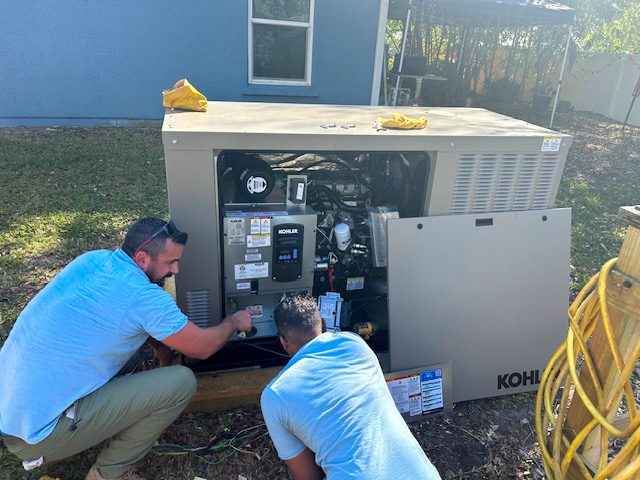 Two technicians in light blue shirts kneel beside an open Kohler generator, inspecting or adjusting internal components in a sunny backyard setting.