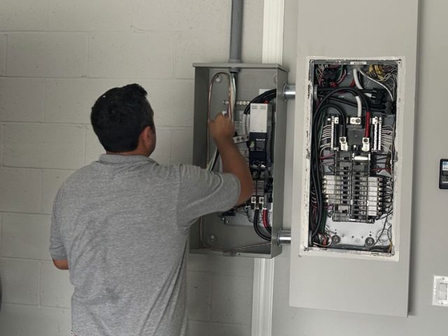 An electrician in Melbourne, FL, is installing and wiring a circuit breaker panel on the wall of a building. The technician, wearing a tool belt, is focused on connecting wires and ensuring the system is properly set up. The open panel reveals a complex arrangement of wires and circuit breakers. The setting appears to be indoors, with a white brick wall in the background. Various tools and equipment are visible, highlighting the professional installation process.