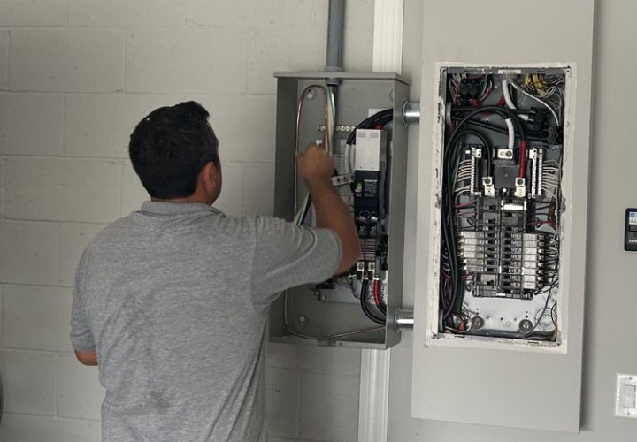 An electrician in Melbourne, FL, is installing and wiring a circuit breaker panel on the wall of a building. The technician, wearing a tool belt, is focused on connecting wires and ensuring the system is properly set up. The open panel reveals a complex arrangement of wires and circuit breakers. The setting appears to be indoors, with a white brick wall in the background. Various tools and equipment are visible, highlighting the professional installation process.