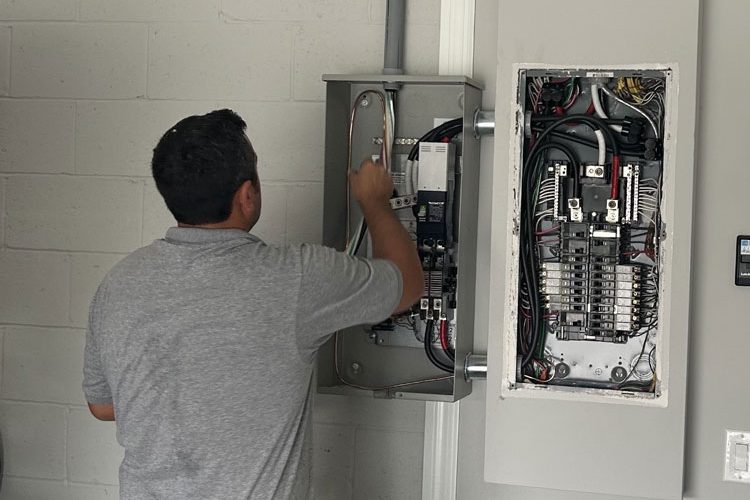 An electrician in Melbourne, FL, is installing and wiring a circuit breaker panel on the wall of a building. The technician, wearing a tool belt, is focused on connecting wires and ensuring the system is properly set up. The open panel reveals a complex arrangement of wires and circuit breakers. The setting appears to be indoors, with a white brick wall in the background. Various tools and equipment are visible, highlighting the professional installation process.