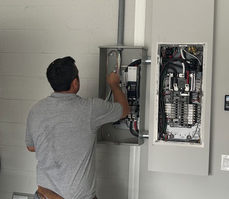 An electrician in Melbourne, FL, is installing and wiring a circuit breaker panel on the wall of a building. The technician, wearing a tool belt, is focused on connecting wires and ensuring the system is properly set up. The open panel reveals a complex arrangement of wires and circuit breakers. The setting appears to be indoors, with a white brick wall in the background. Various tools and equipment are visible, highlighting the professional installation process.
