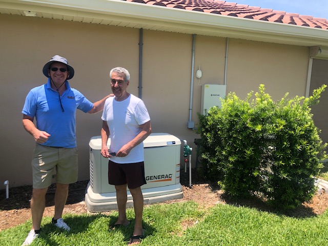 A homeowner stands with an electrical contractor beside a newly installed Generac generator outside a beige house. Both individuals are smiling, indicating satisfaction with the installation. The generator is positioned on a concrete pad next to the house, with utility connections visible. The surrounding area is landscaped with green grass and shrubs, and the house has a tiled roof, highlighting the residential setting.