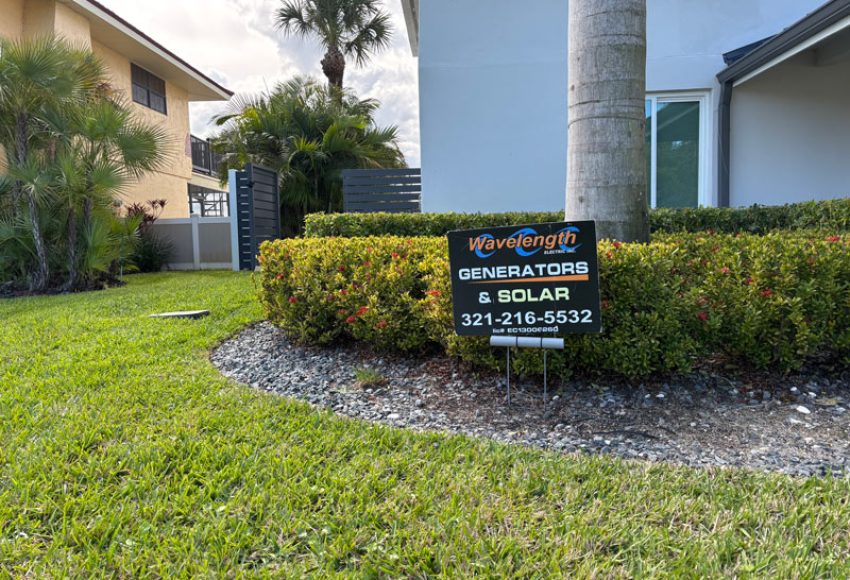 A black yard sign reading "Wavelength Generators & Solar" with contact information, placed in a well-maintained lawn surrounded by shrubs and palm trees near a residential building.