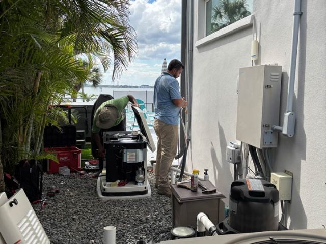 Two technicians work on a generator and electrical equipment in a narrow outdoor space beside a white stucco wall, with tools, hoses, and HVAC units nearby, under a partly cloudy sky.