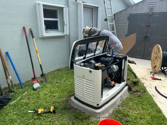 A technician in Melbourne, FL, works on installing a backup generator outside a house. The generator's cover is open, revealing its internal components. Various tools and equipment are scattered around, including shovels, a power drill, and a spool of cable. The installation site is next to a gray house with open windows and a shed in the background.