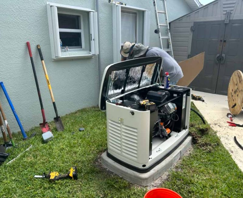 A technician in Melbourne, FL, works on installing a backup generator outside a house. The generator's cover is open, revealing its internal components. Various tools and equipment are scattered around, including shovels, a power drill, and a spool of cable. The installation site is next to a gray house with open windows and a shed in the background.