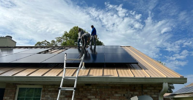 Technicians in Melbourne, FL, are installing solar panels on the roof of a brick house. The panels are arranged in rows, and the workers are positioned on the roof, securing the panels. A ladder is propped against the house, and a satellite dish is visible on the roof. The sky is partly cloudy, providing a bright backdrop for the installation process.