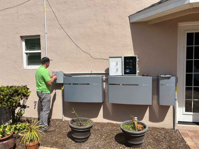 An electrical technician in Melbourne, FL, works on installing and connecting multiple control panels on the exterior wall of a house. The technician, wearing a green shirt, is seen adjusting one of the panels. The setup includes various control boxes and wiring conduits. The area around the installation is landscaped with potted plants and a small garden, adding a touch of greenery to the scene. The background includes a small window and a door leading into the house.