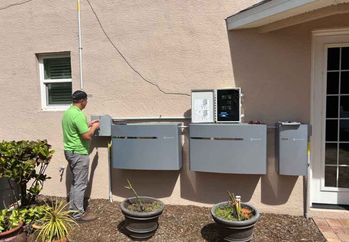 An electrical technician in Melbourne, FL, works on installing and connecting multiple control panels on the exterior wall of a house. The technician, wearing a green shirt, is seen adjusting one of the panels. The setup includes various control boxes and wiring conduits. The area around the installation is landscaped with potted plants and a small garden, adding a touch of greenery to the scene. The background includes a small window and a door leading into the house.