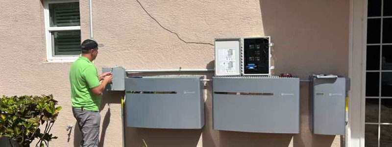 An electrical technician in Melbourne, FL, works on installing and connecting multiple control panels on the exterior wall of a house. The technician, wearing a green shirt, is seen adjusting one of the panels. The setup includes various control boxes and wiring conduits. The area around the installation is landscaped with potted plants and a small garden, adding a touch of greenery to the scene. The background includes a small window and a door leading into the house.