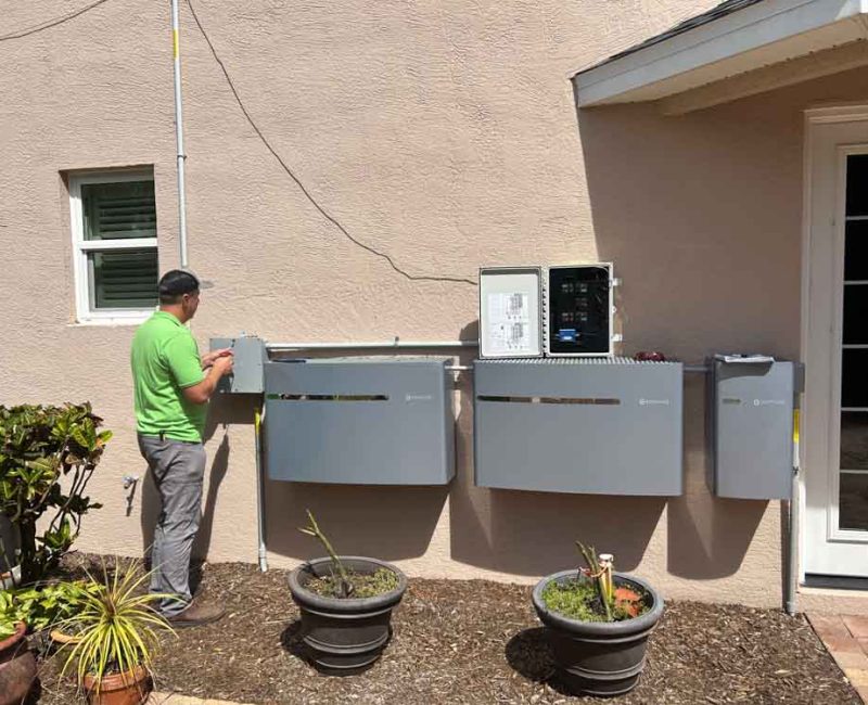 An electrical technician in Melbourne, FL, works on installing and connecting multiple control panels on the exterior wall of a house. The technician, wearing a green shirt, is seen adjusting one of the panels. The setup includes various control boxes and wiring conduits. The area around the installation is landscaped with potted plants and a small garden, adding a touch of greenery to the scene. The background includes a small window and a door leading into the house.