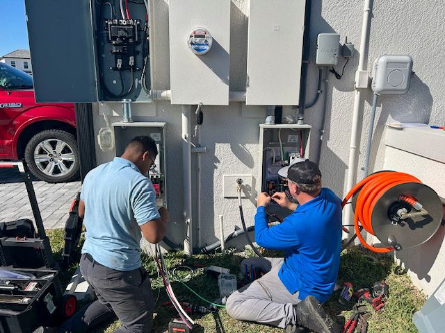 Two electricians work on an outdoor electrical panel system, one kneeling and one standing, using tools and cables, with a red vehicle and coiled orange extension cord visible in the background.